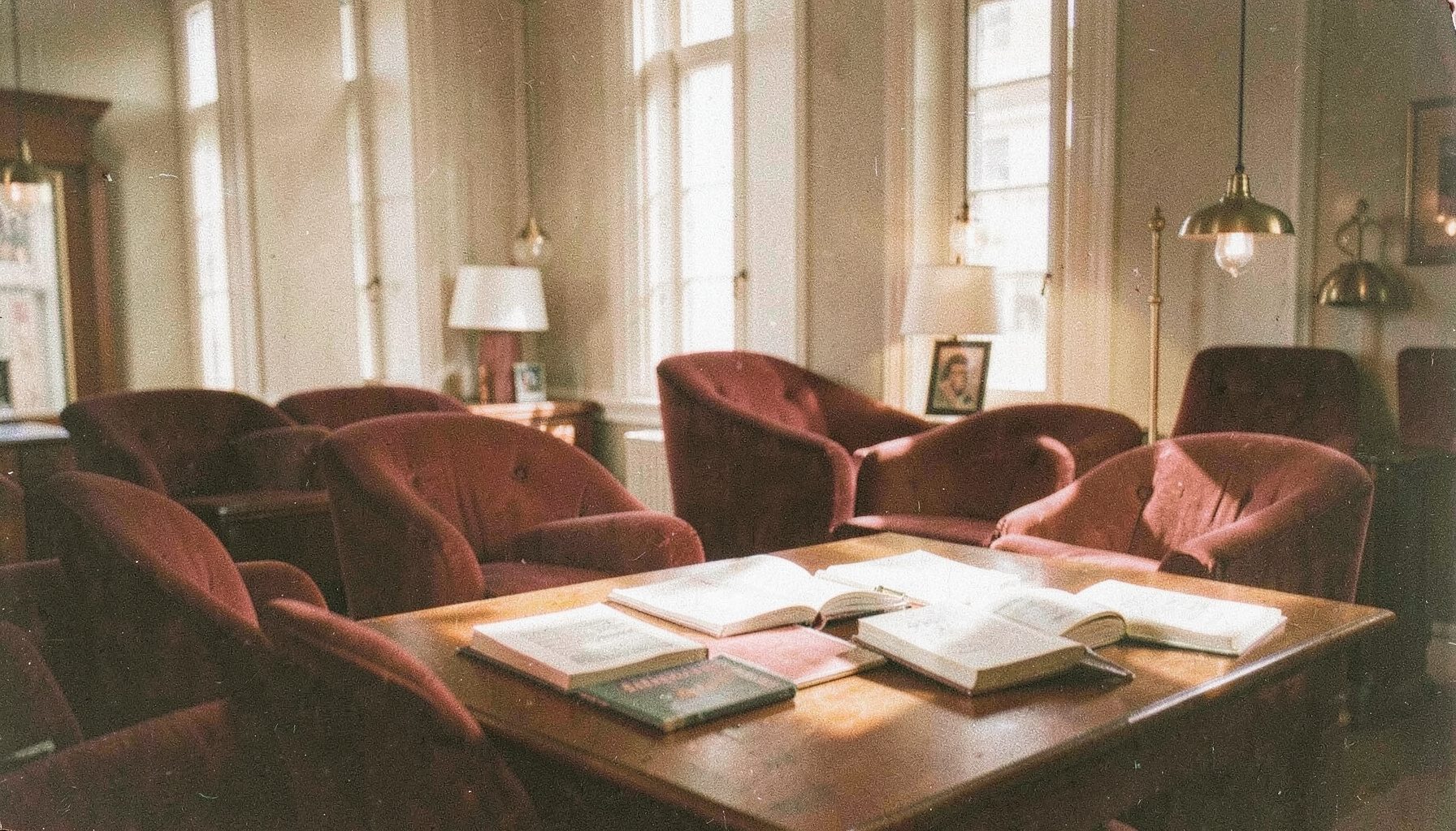 A grainy analog photograph of a sunlit reading room with warm burgundy upholstered chairs, brass-toned lamp fixtures, and soft afternoon light falling across open books on a wooden table