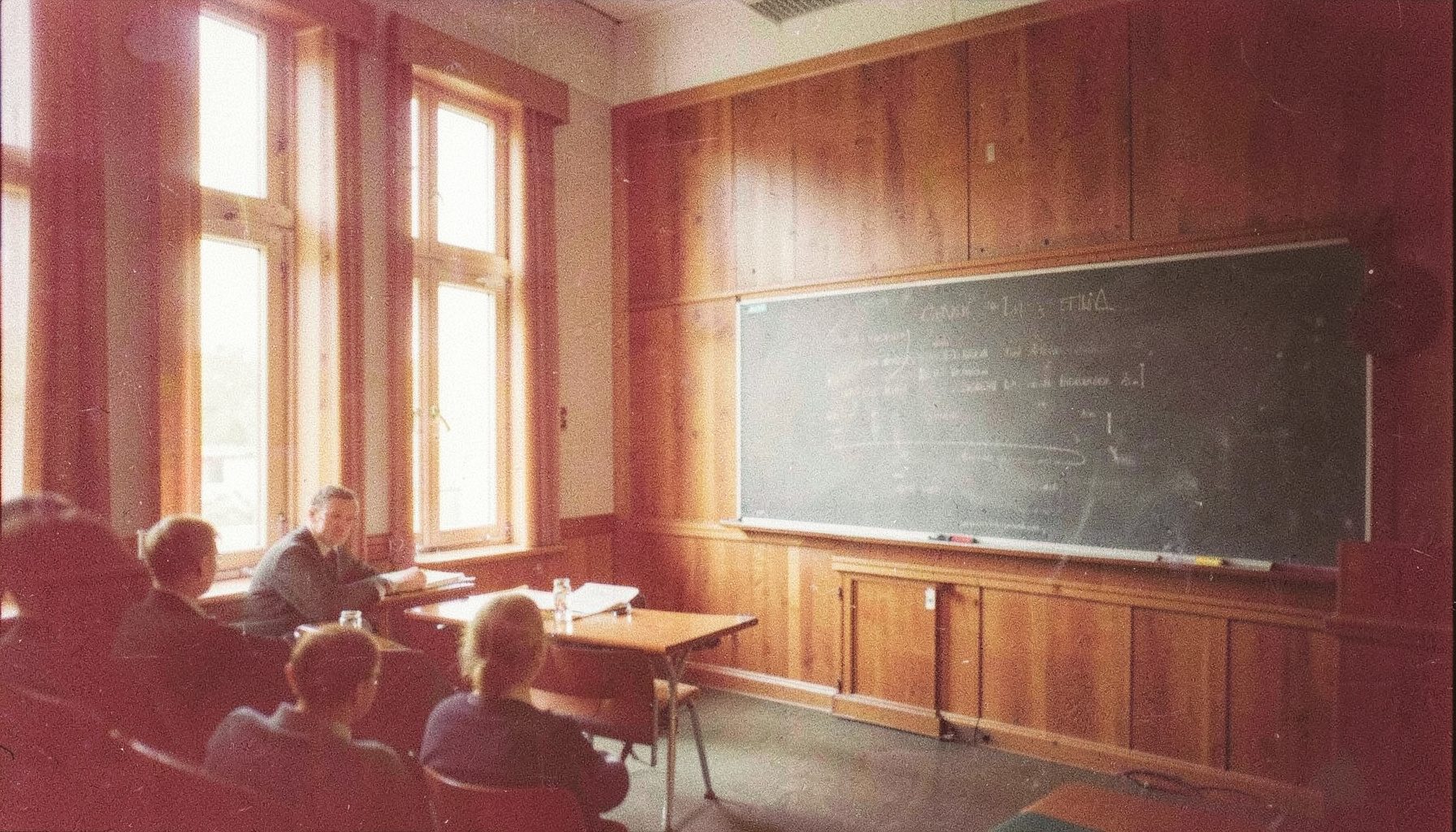 A grainy analog photograph of a seminar room with a chalkboard, warm wood paneling, and soft daylight streaming through tall windows