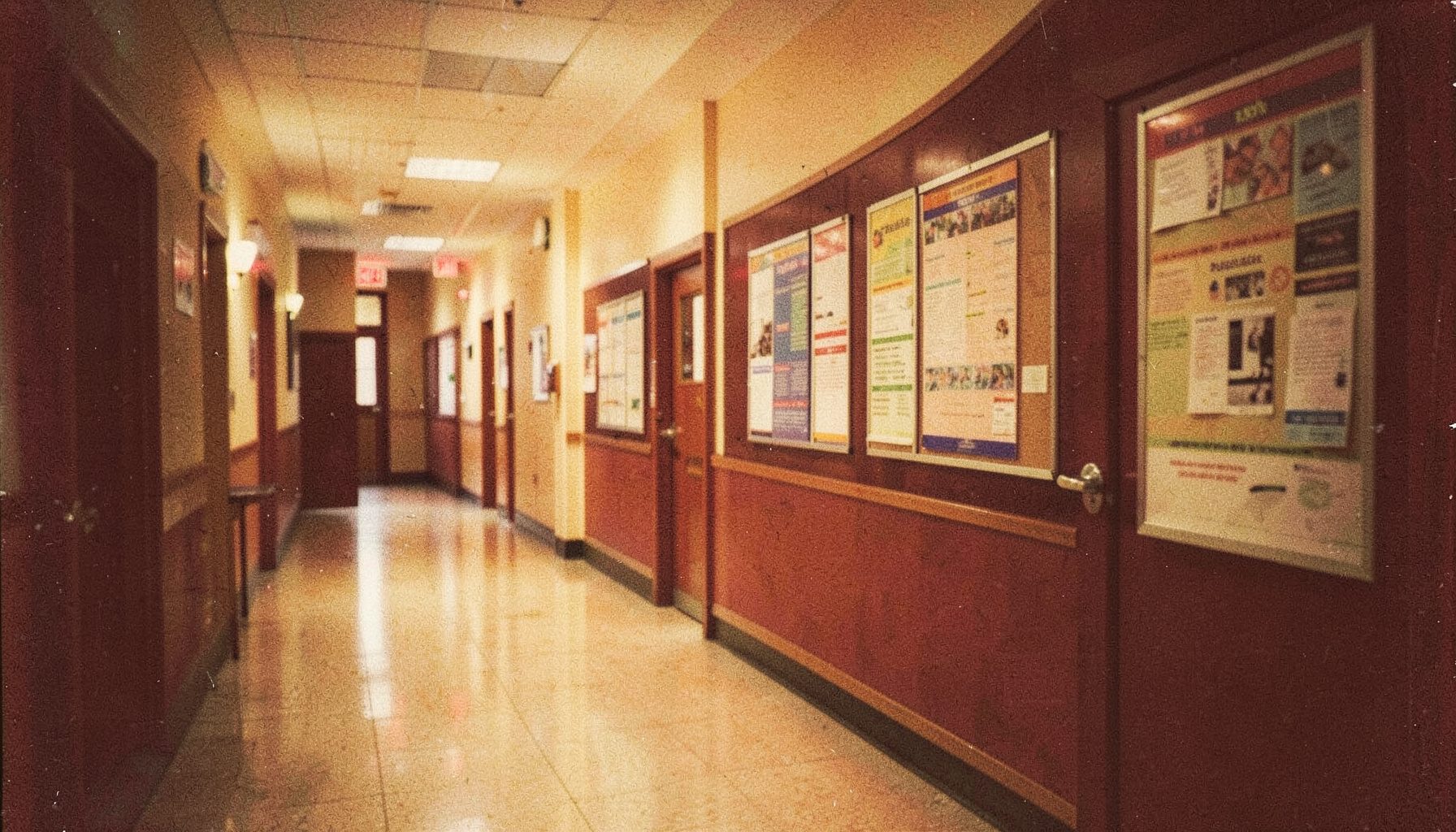 A grainy film photograph of a university hallway with bulletin boards, warm golden light, and rich burgundy accents