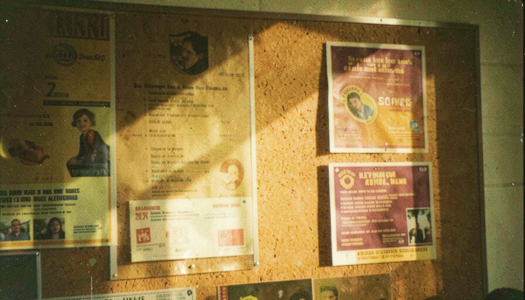 A grainy analog photograph of a campus bulletin board with warm gold and burgundy flyers, soft afternoon light casting deep shadows across layered event announcements