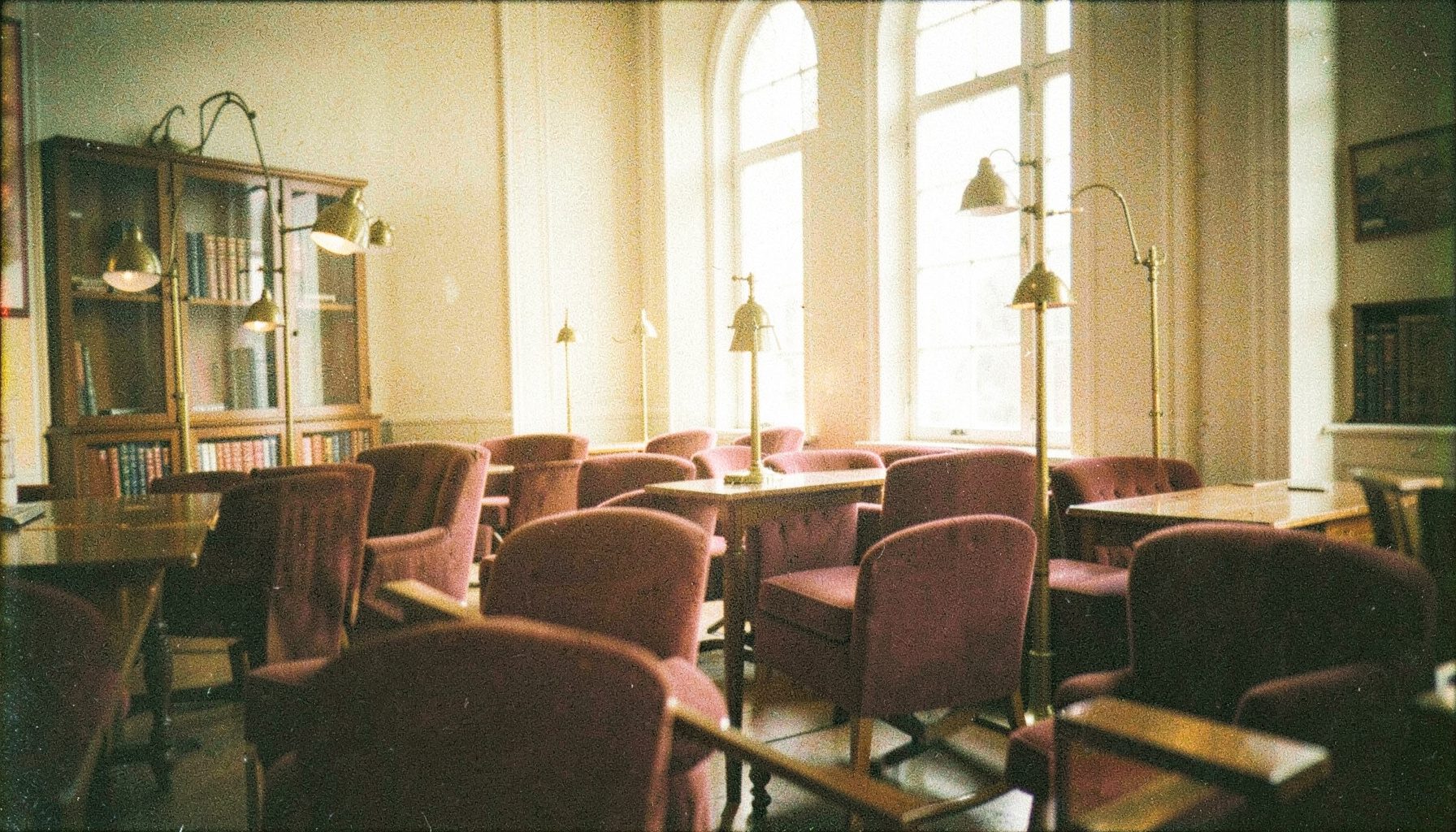 A grainy analog photograph of a sunlit reading room with warm burgundy upholstered chairs and brass-toned lamp fixtures, conveying a quiet academic atmosphere.