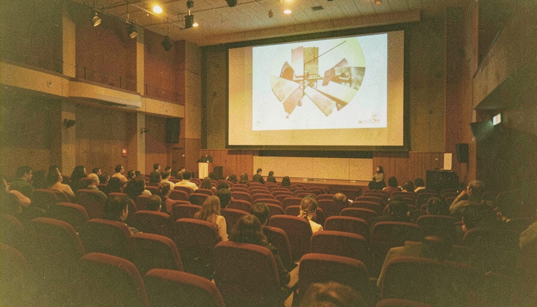 A grainy film photograph of a lecture hall interior with rich burgundy seating, warm amber lighting, and a large projection screen at the front, conveying a quiet academic atmosphere.